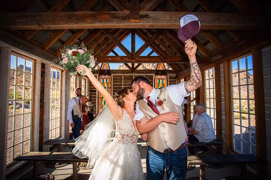 Wedding couple celebrating in a rustic chapel at Nelson Ghost Town, bride lifting her bouquet and groom raising his hat in warm golden light